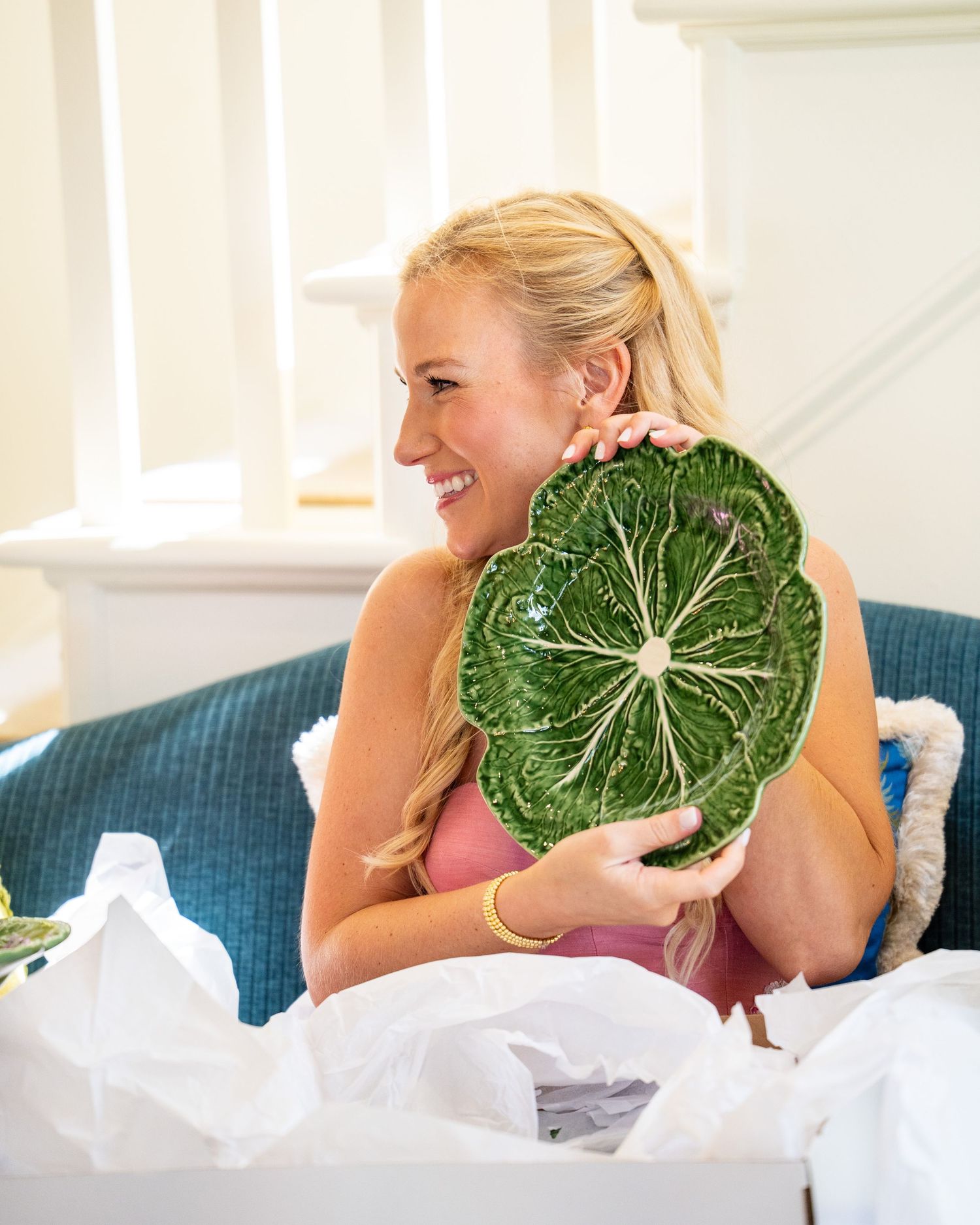 Smiling blonde woman holding a decorative green cabbage leaf plate while sitting on a teal sofa.