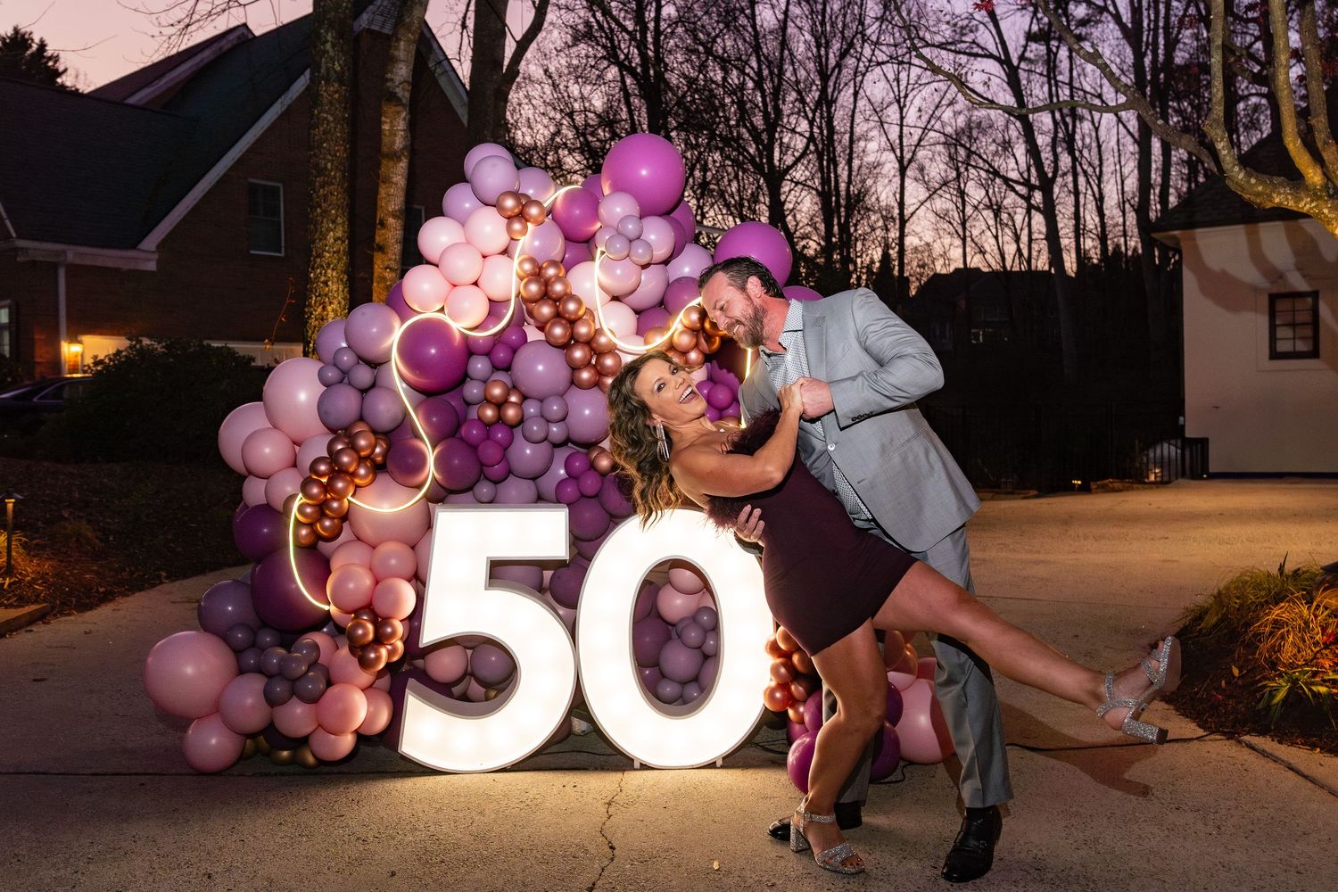 Couple posing romantically in front of a glowing 50 sign with purple balloon arch at a 50th birthday celebration.