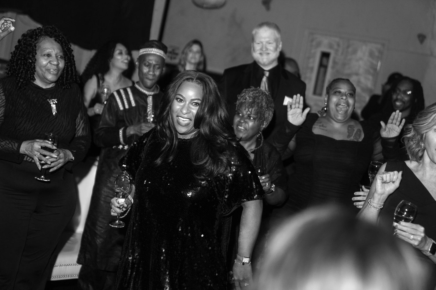 Black and white photo of a woman in a sequined dress holding a drink, surrounded by a group of smiling people at a party.