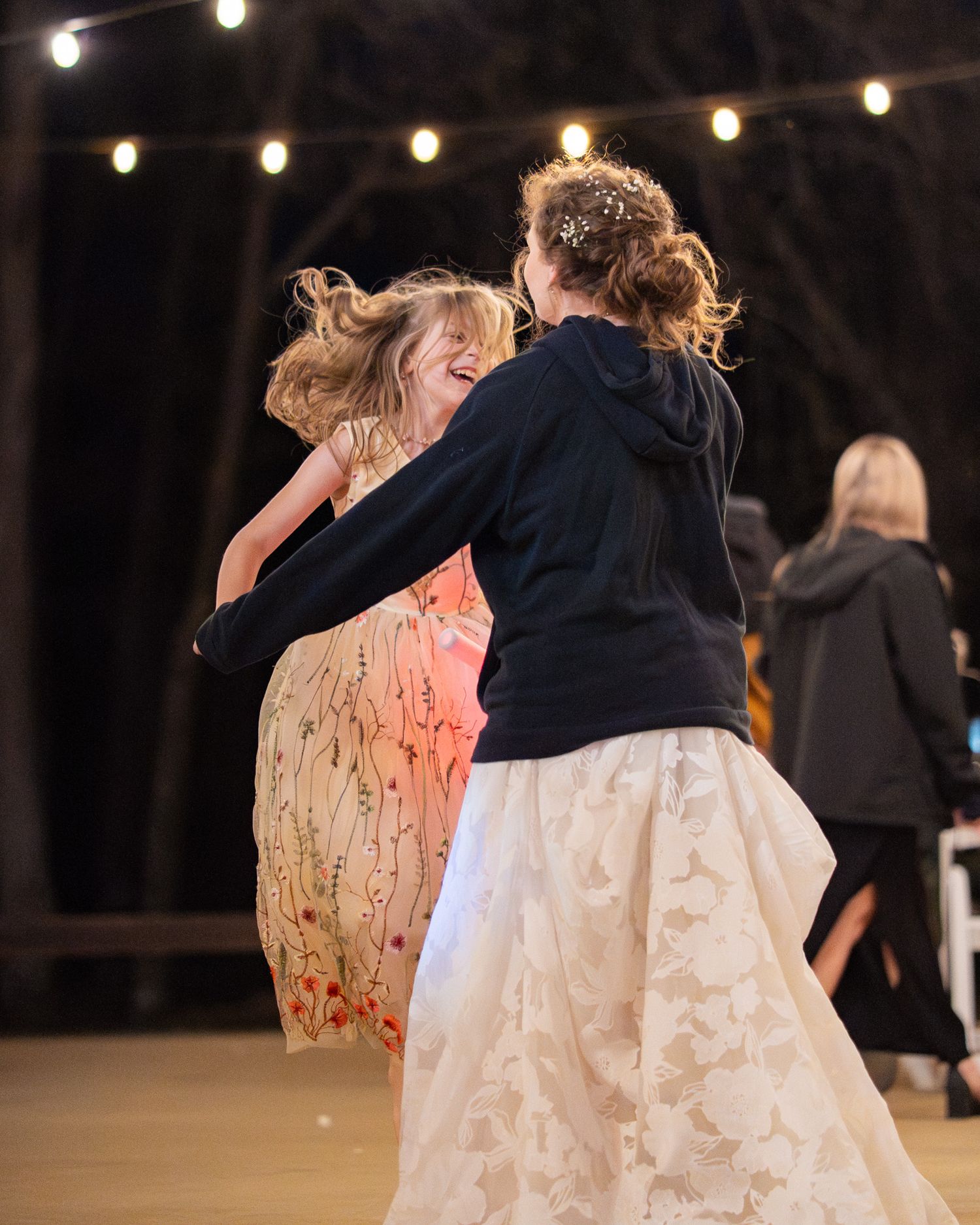 Two women dancing joyfully outdoors at a wedding reception under string lights at night.