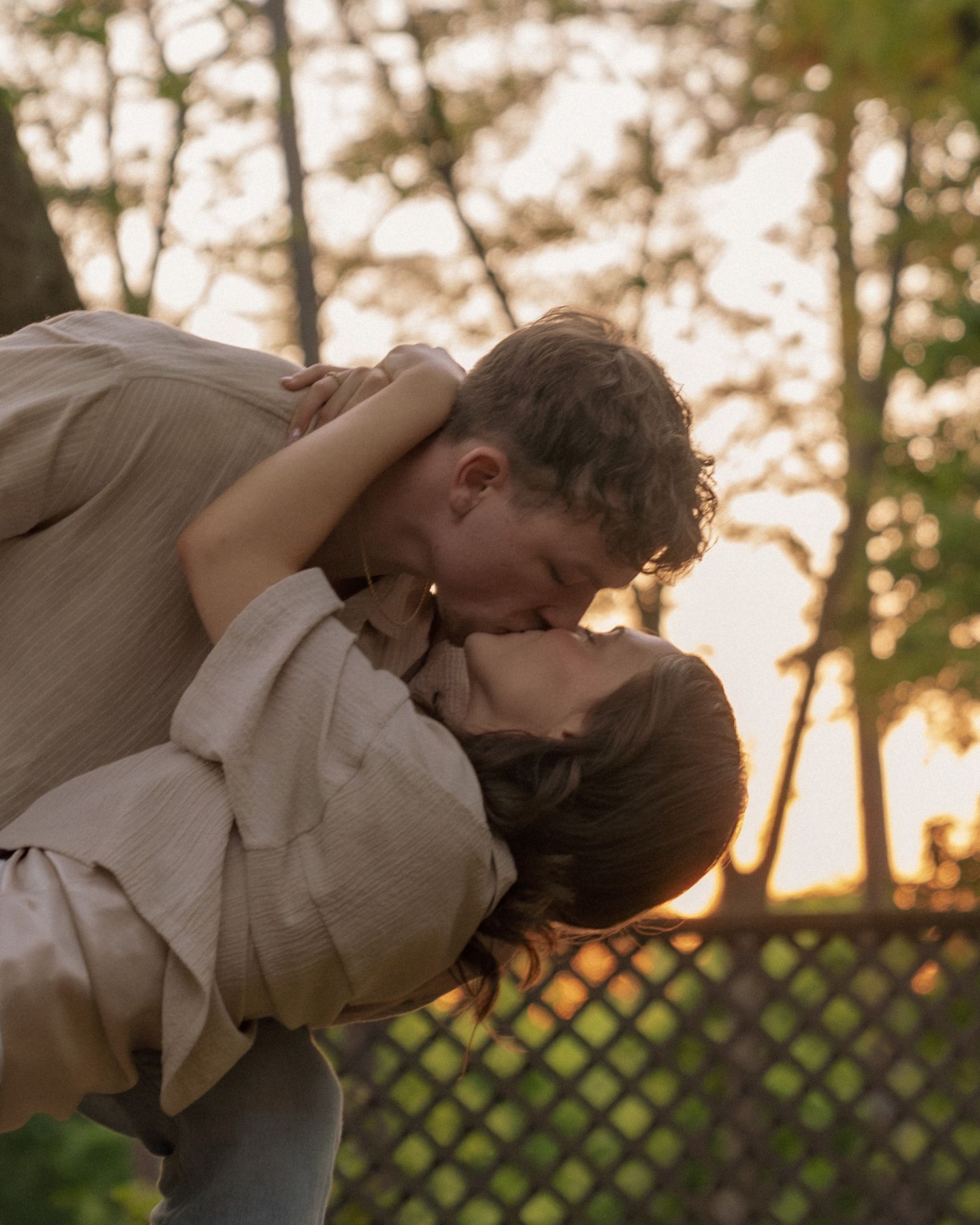 Couple sharing a romantic kiss at golden hour outdoors, embracing near trees with warm sunset light.