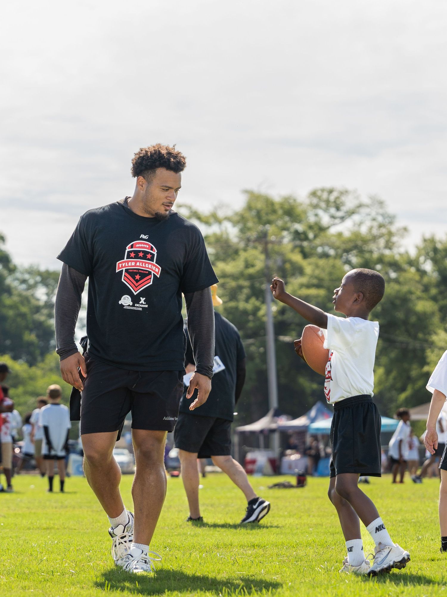 Tall athlete in black team shirt mentors young child holding football on green field at youth sports camp.