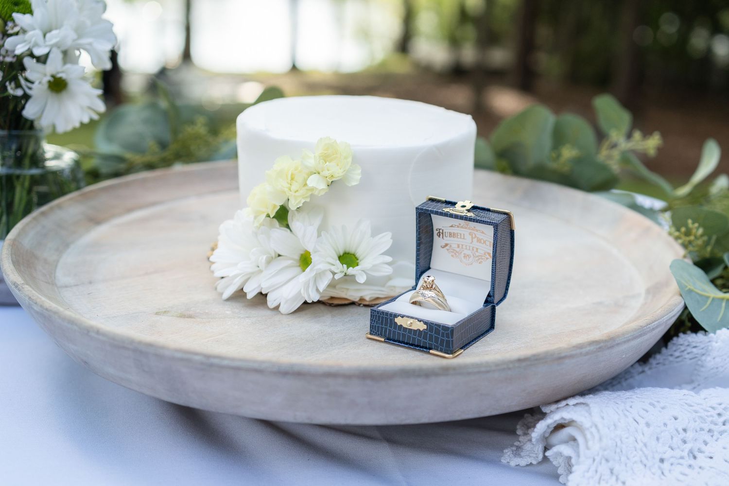 White wedding cake with daisies and gold rings in a navy box on a wooden tray at an outdoor wedding.