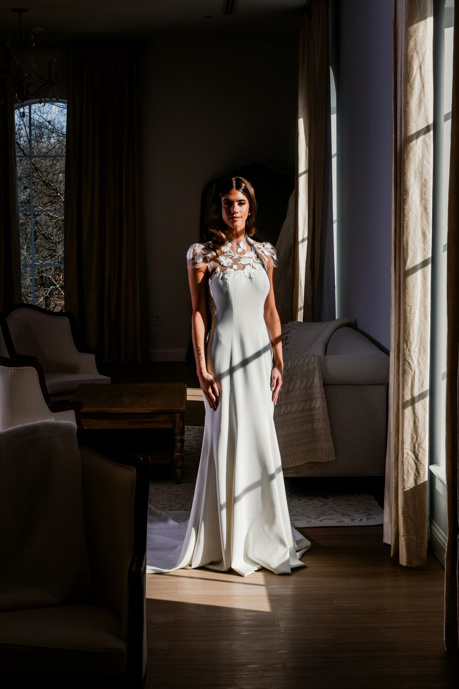 Bride in elegant white gown with floral shoulder detail stands in sunlit room with large windows.