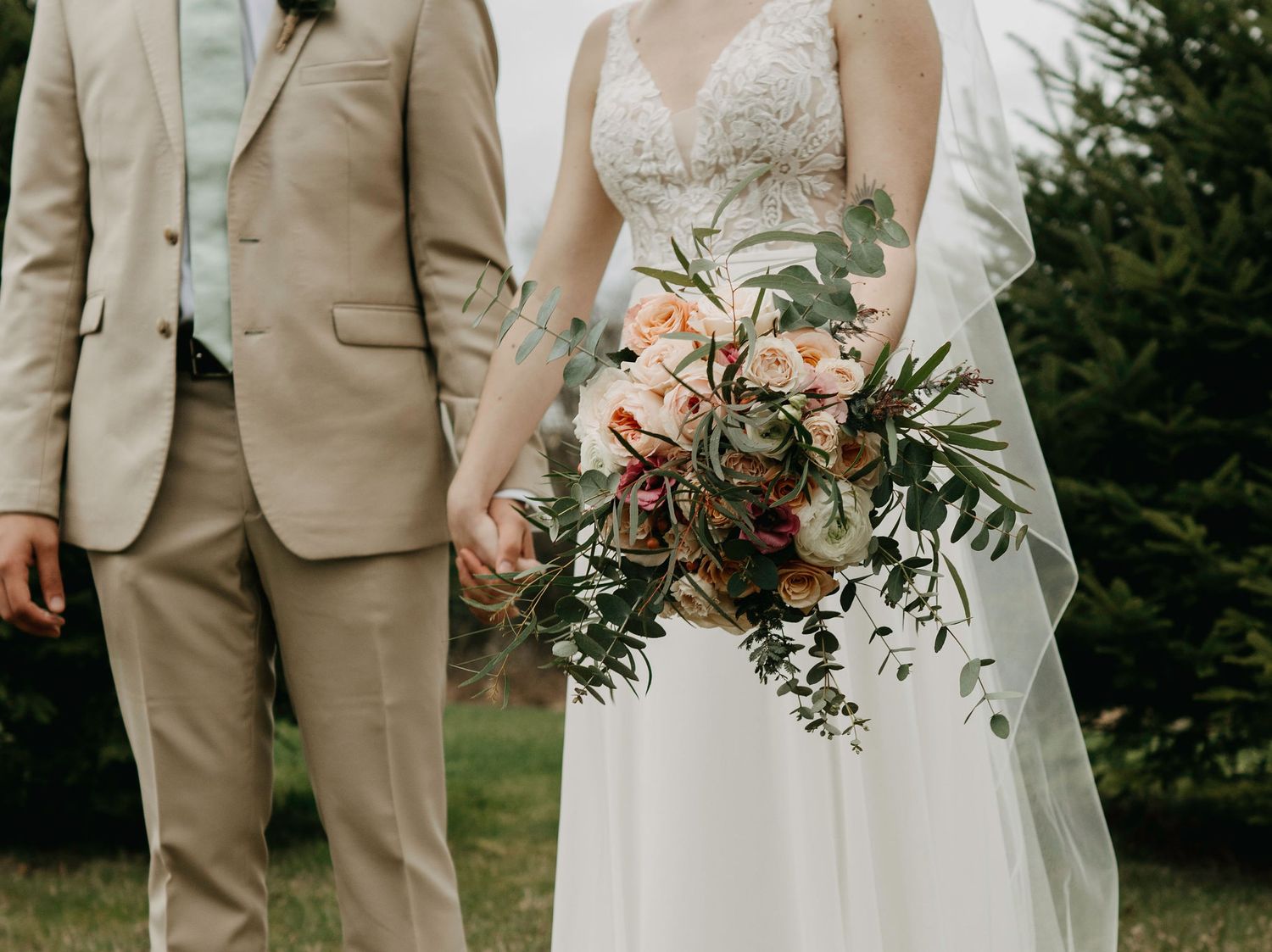 Bride holding a lush bouquet of peach roses and greenery beside groom in a tan suit outdoors.