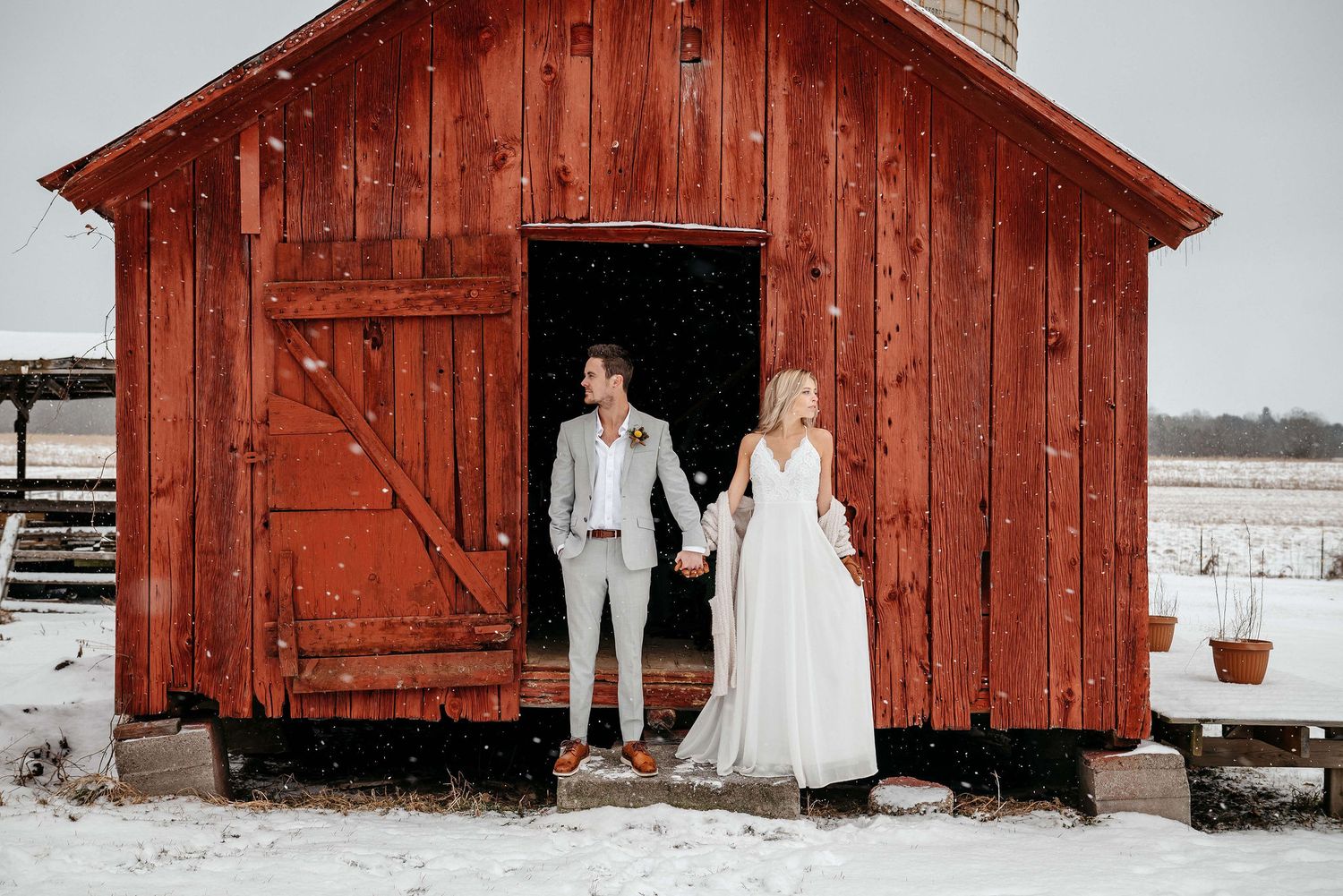 Bride and groom holding hands in front of a rustic red barn in a snowy winter wedding setting.