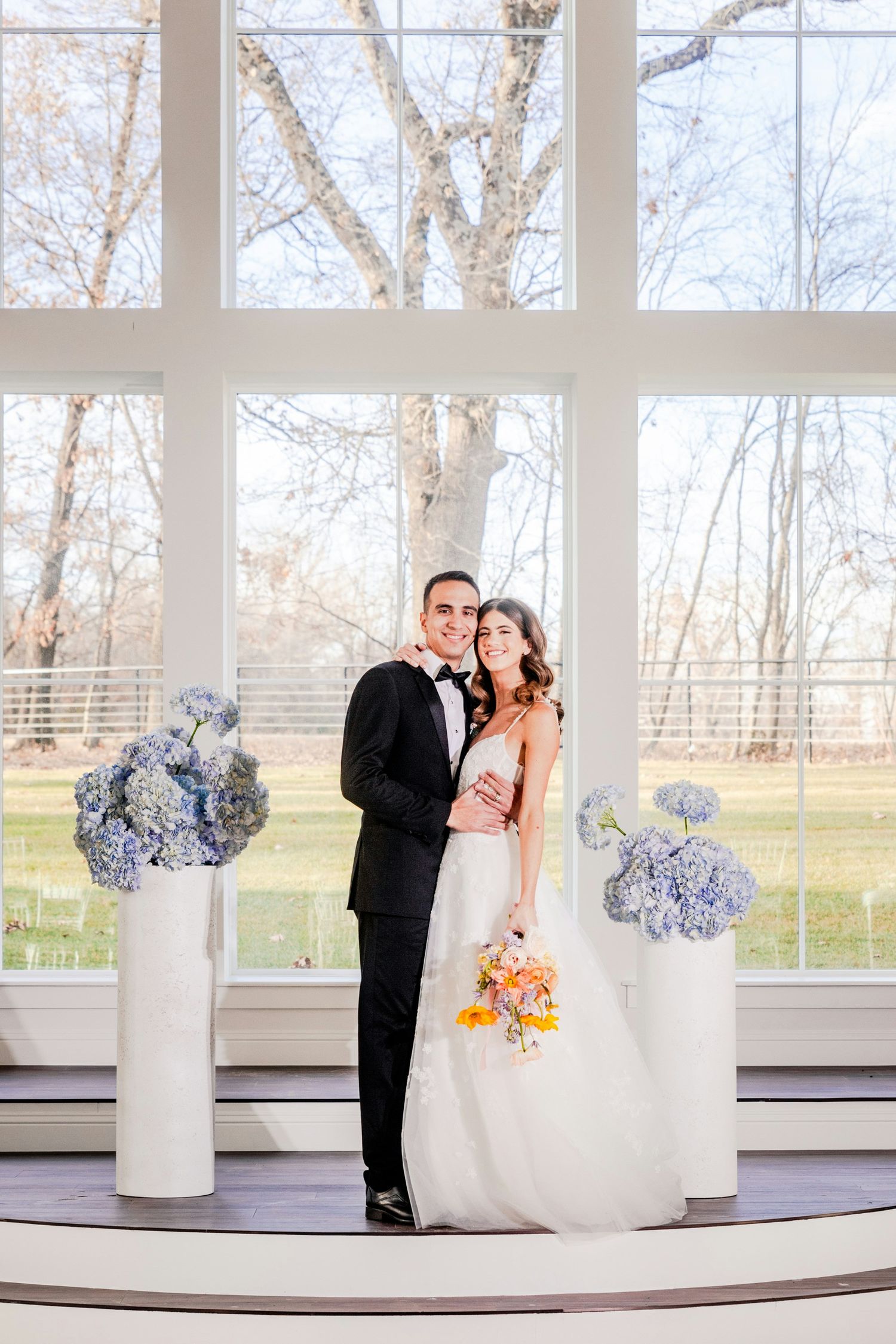Bride and groom pose between white pedestals with blue hydrangeas before large windows at elegant wedding venue.