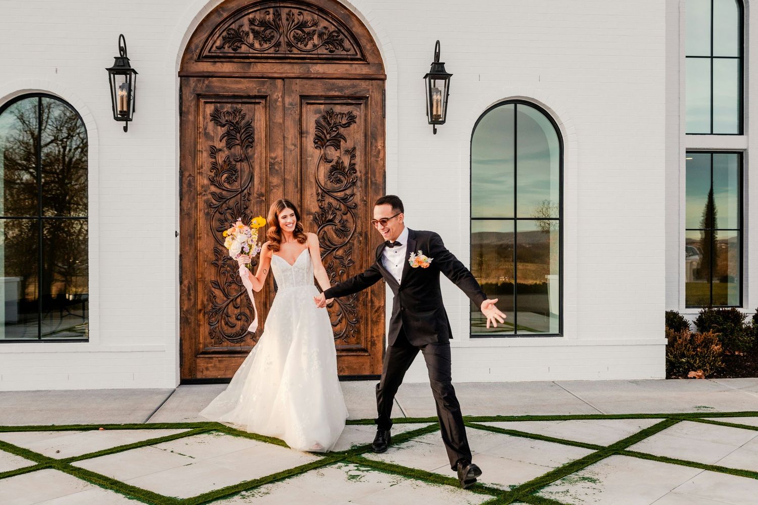 Joyful bride and groom dancing outside elegant white wedding venue with ornate wooden doors and arched windows.