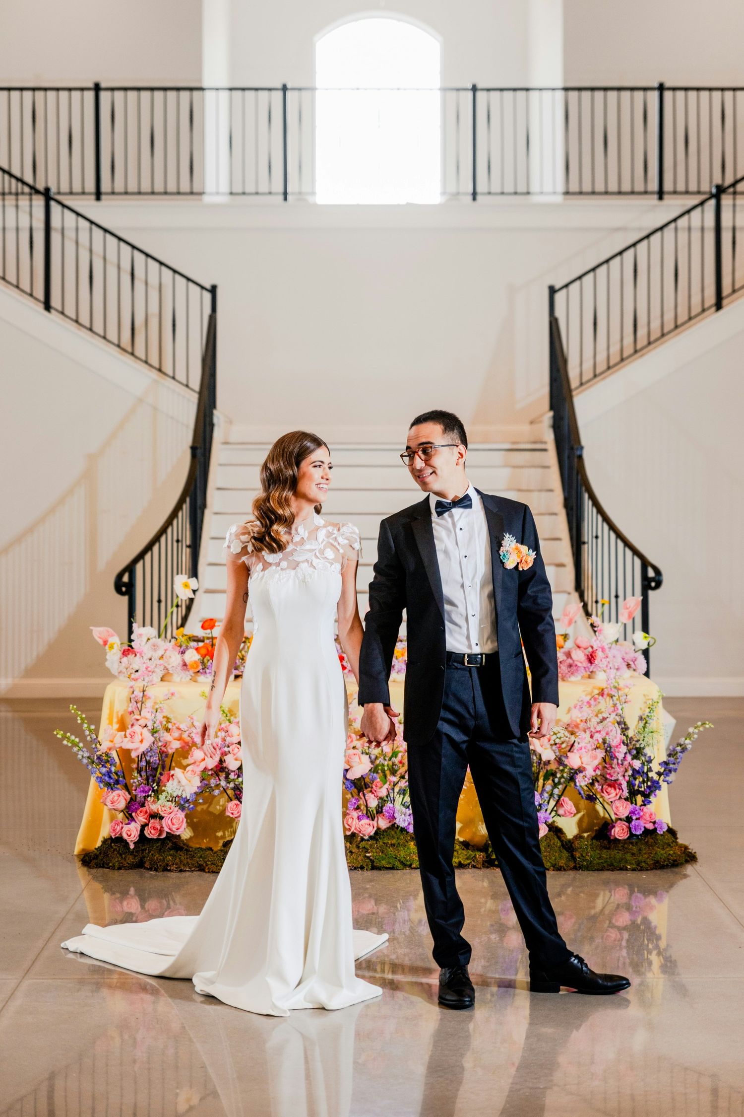 Bride and groom hold hands on marble floor before grand staircase adorned with colorful floral arrangements.