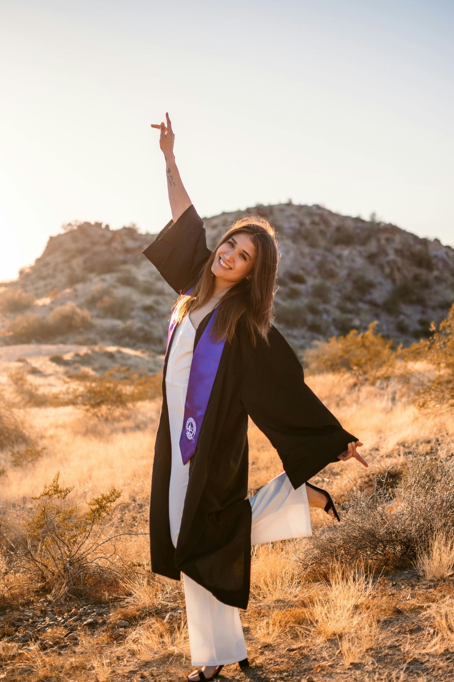 Smiling graduate in black cap and gown celebrating outdoors in desert landscape at sunset.