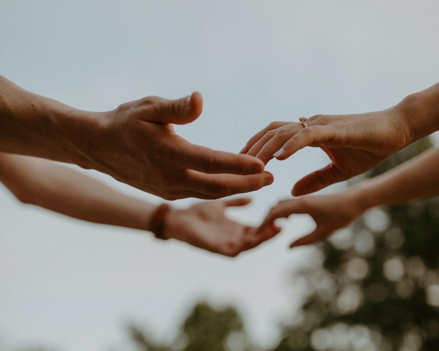 Two hands reaching toward each other against a soft sky background with an engagement ring visible.