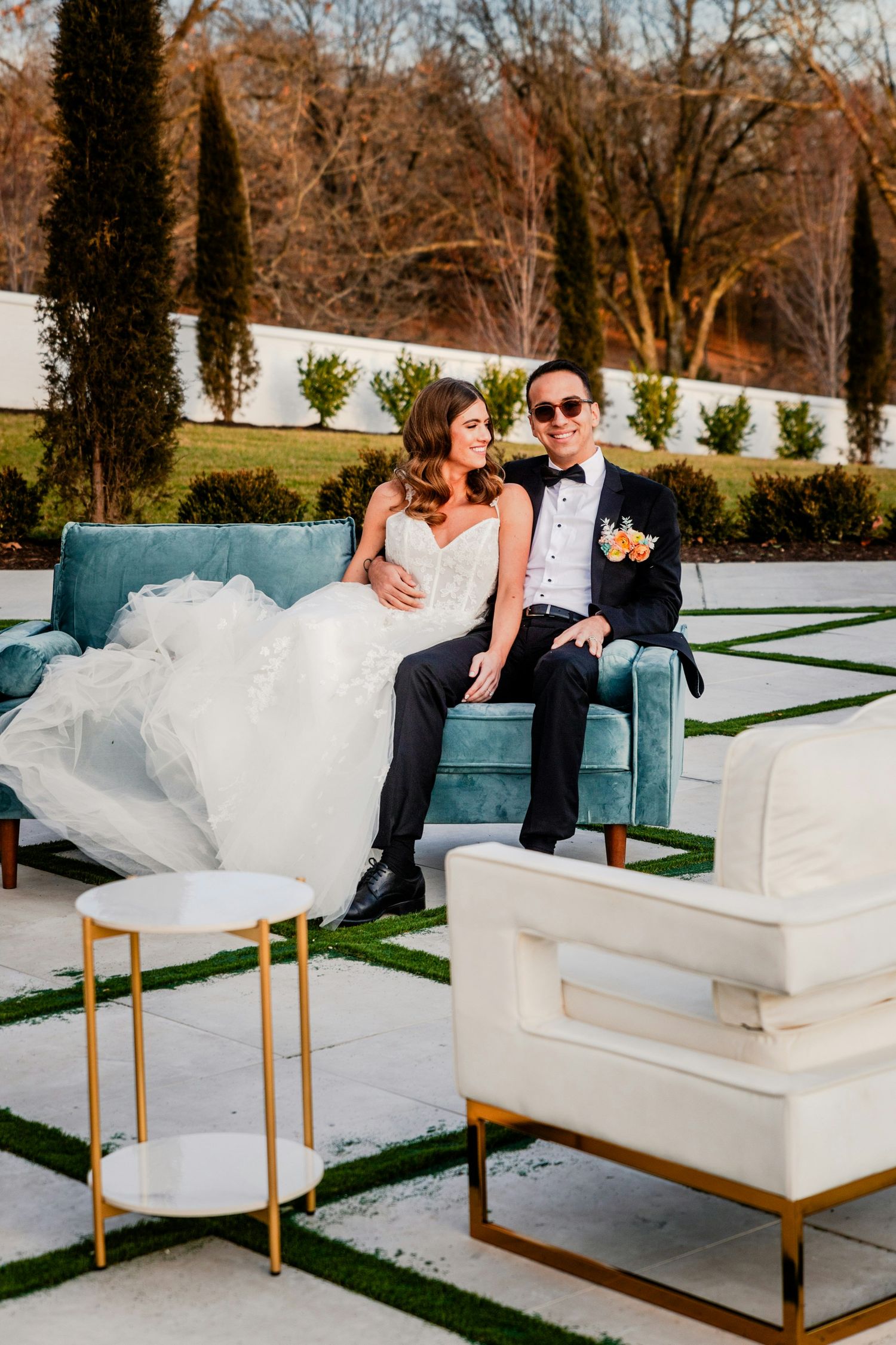 Bride and groom sit closely on teal velvet sofa in elegant outdoor garden setting with cypress trees.