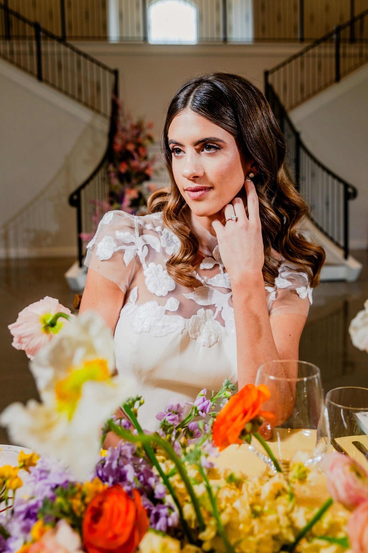 Bride in white floral gown sits at colorful flower-adorned reception table in elegant venue with grand staircase.