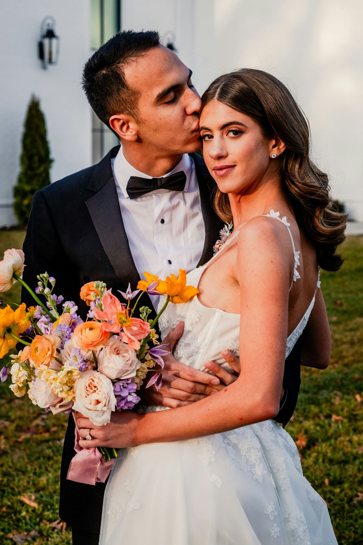 Groom in black tuxedo kissing bride on temple while she holds colorful spring wedding bouquet outdoors.