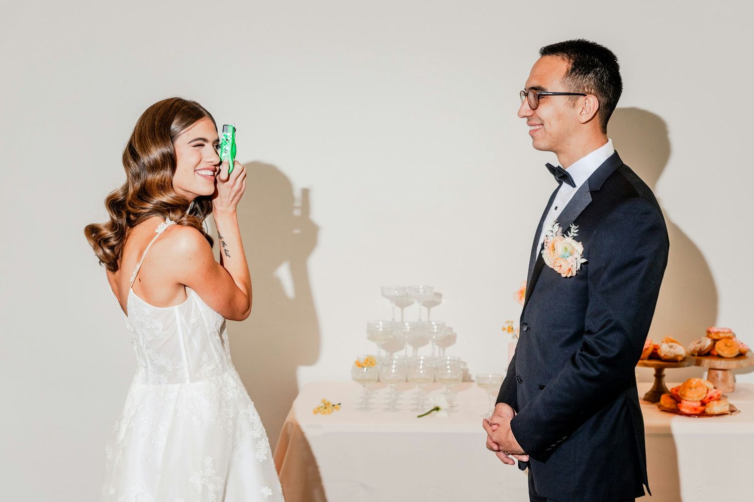 Smiling bride photographs groom with green can near champagne tower at elegant wedding reception against neutral background.