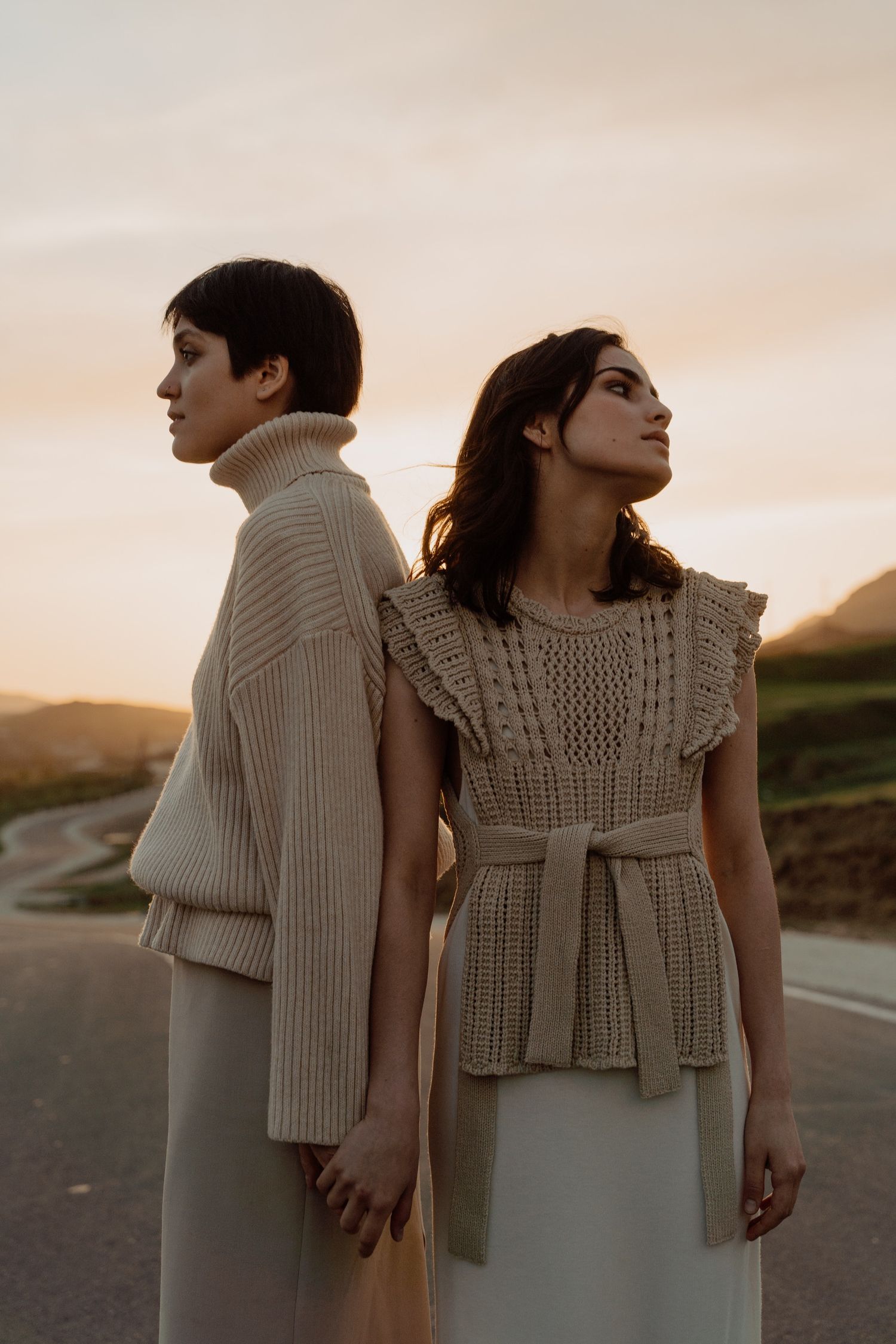 Two women in neutral knitwear stand back to back on a road at golden hour sunset.