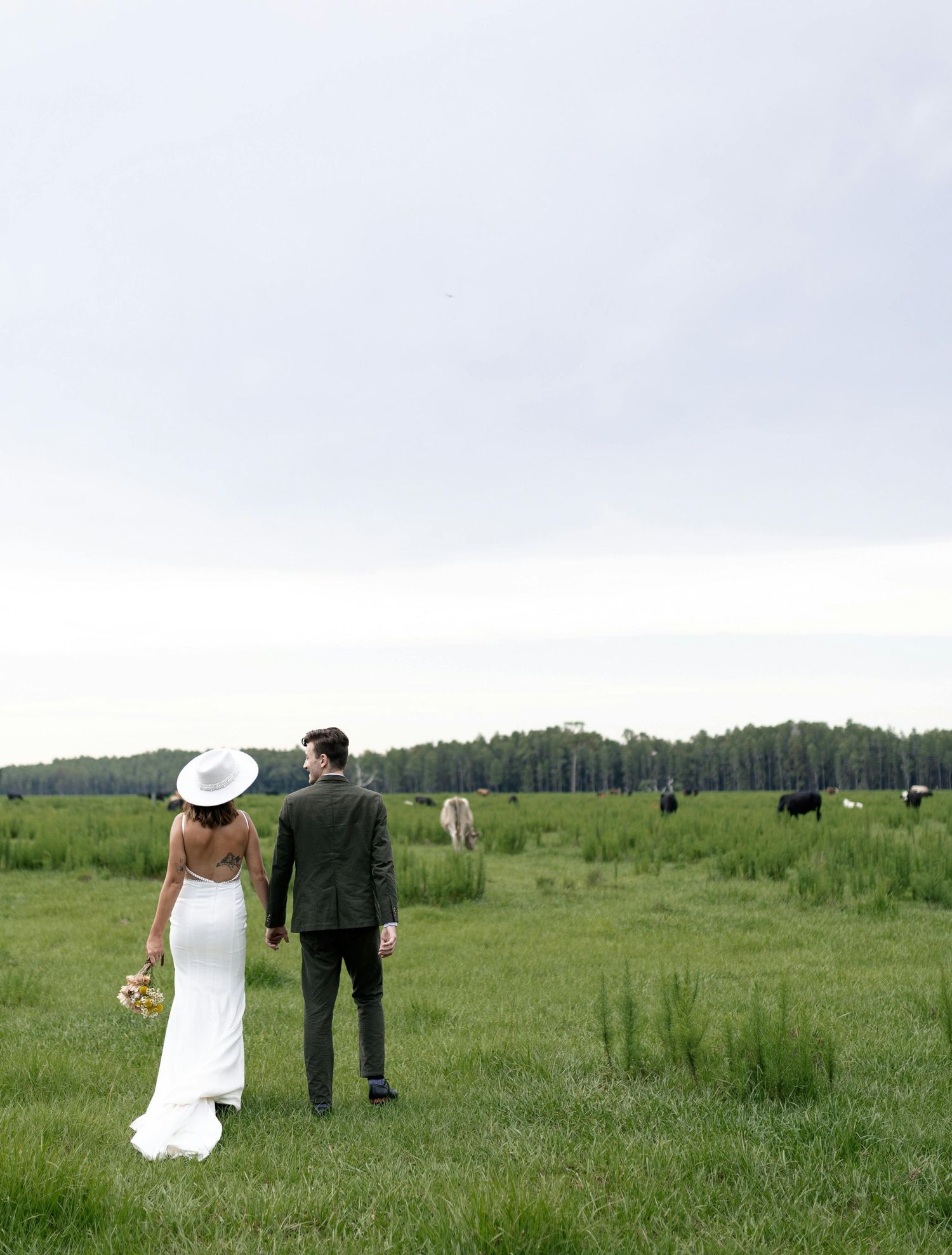 Bride and groom standing in green meadow with grazing cattle and forest backdrop.