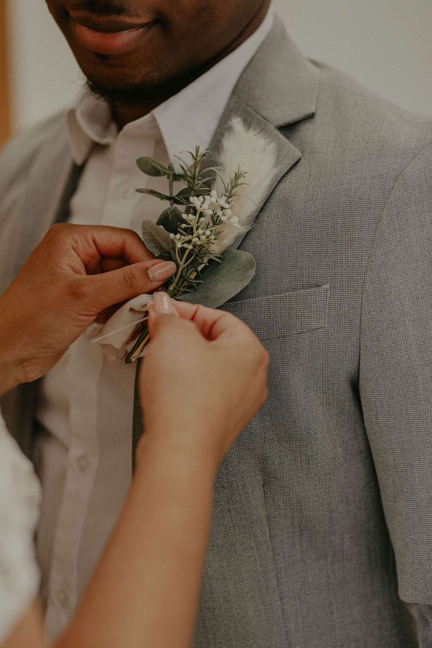 Hands pinning a greenery and white floral boutonniere onto a groom's gray suit jacket.
