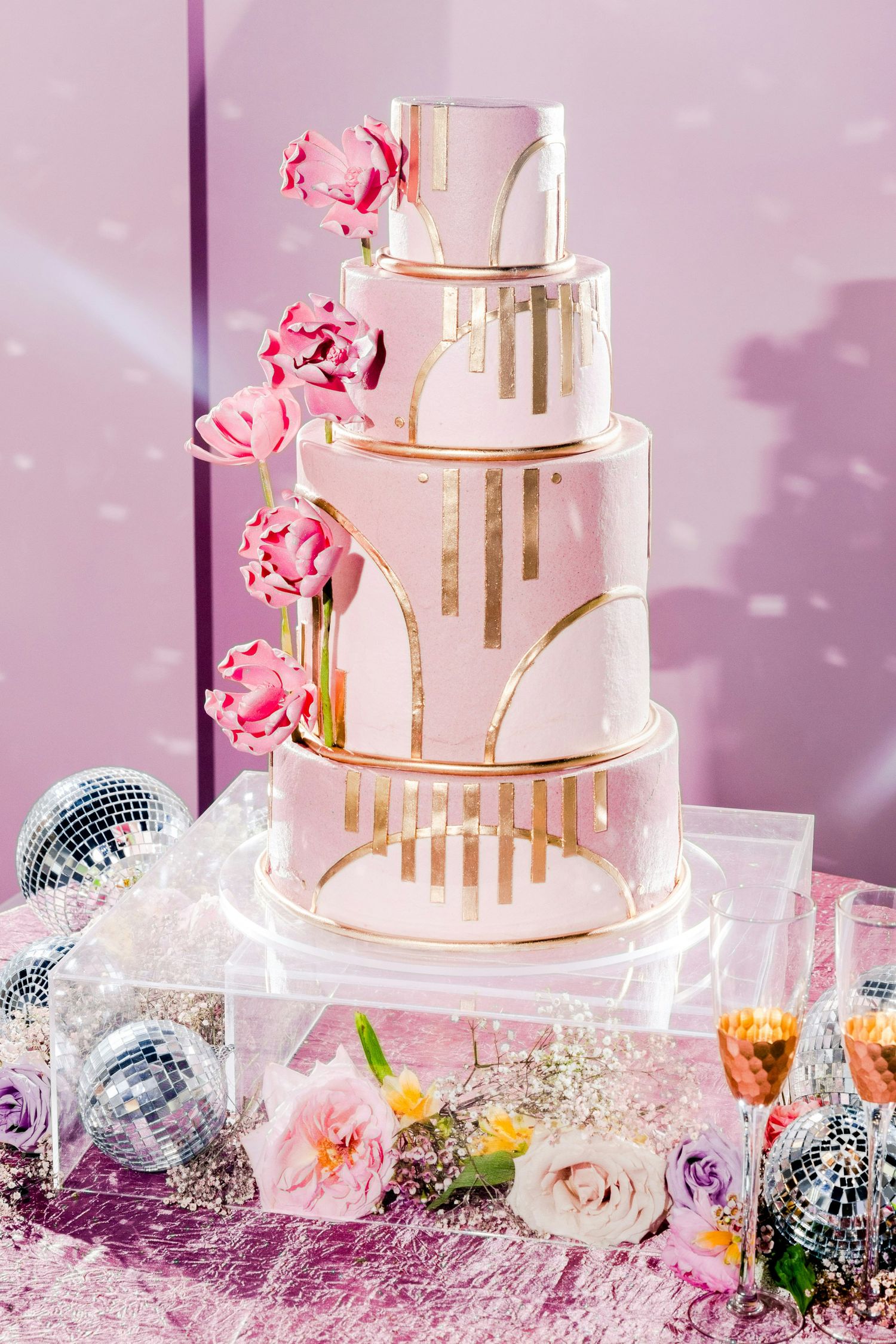 Elegant four-tier pink and gold wedding cake adorned with pink tulips and Art Deco gold details on a acrylic stand.