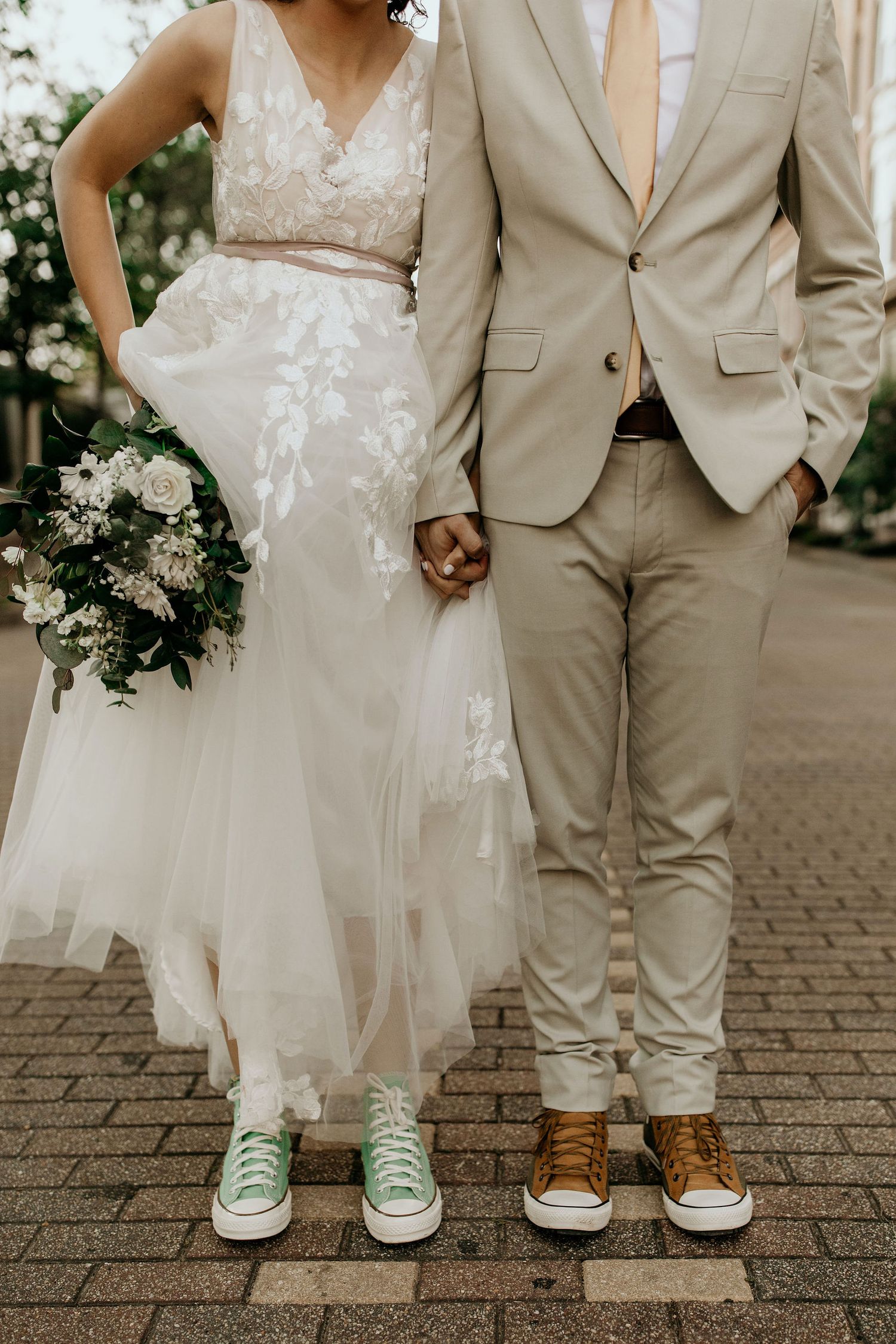 Wedding couple holding hands wearing casual sneakers, bride in floral lace gown holding white bouquet, groom in beige suit.