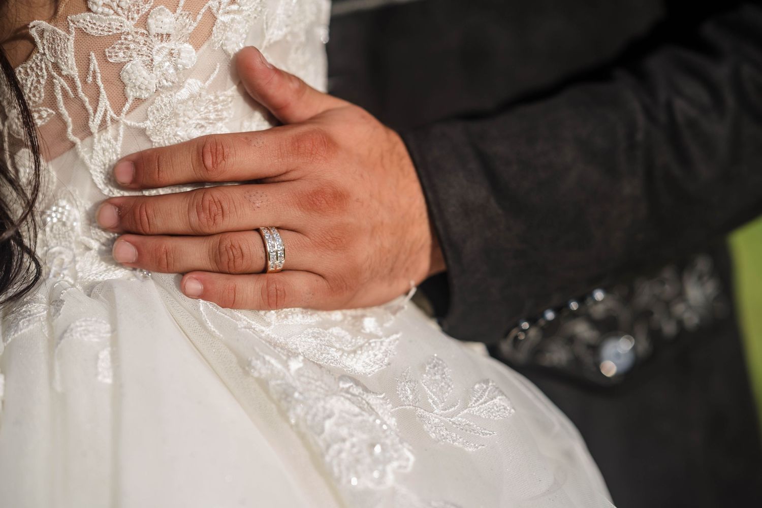 Close-up of groom's hand with wedding ring resting on bride's white lace wedding dress.