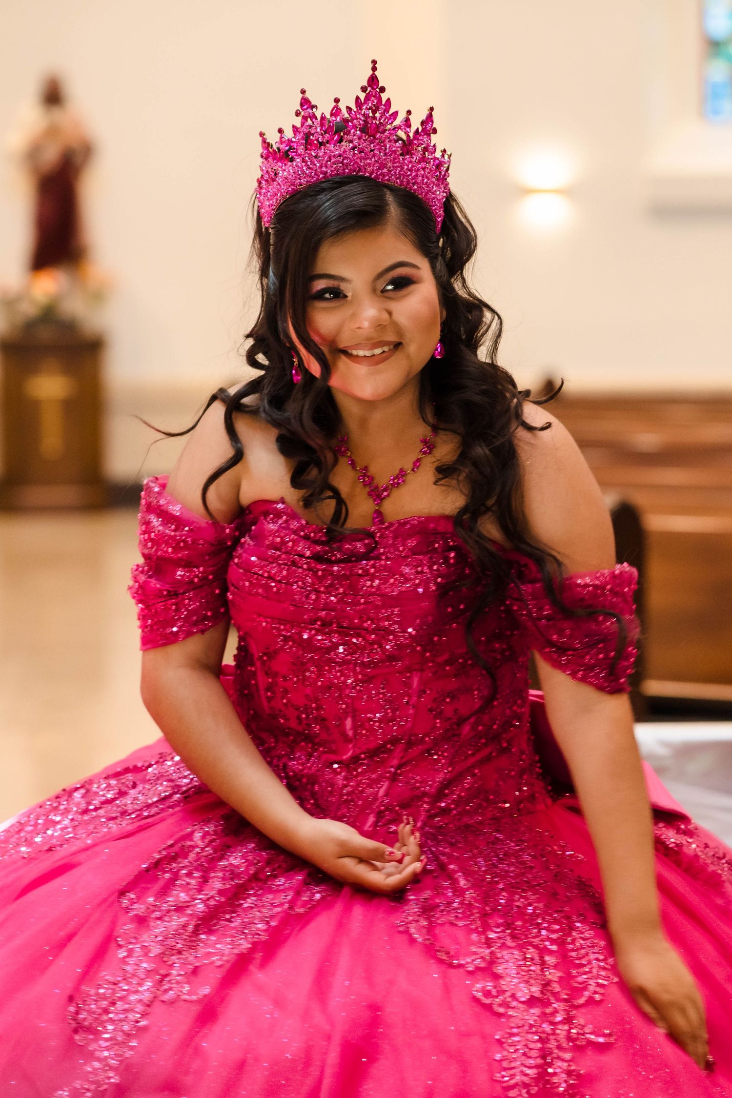 Smiling quinceañera girl in hot pink sequined gown and crown sitting in a church.