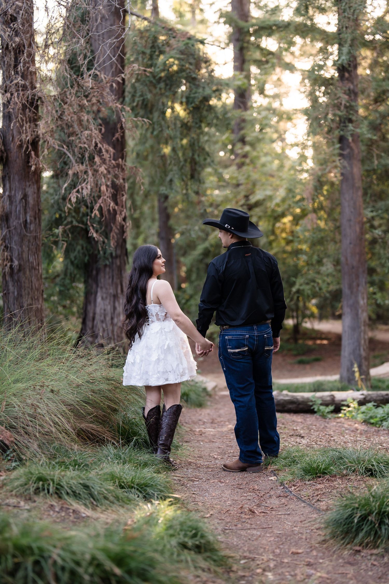 Couple walking hand in hand on a forest path, man in black cowboy hat, woman in white dress and boots.