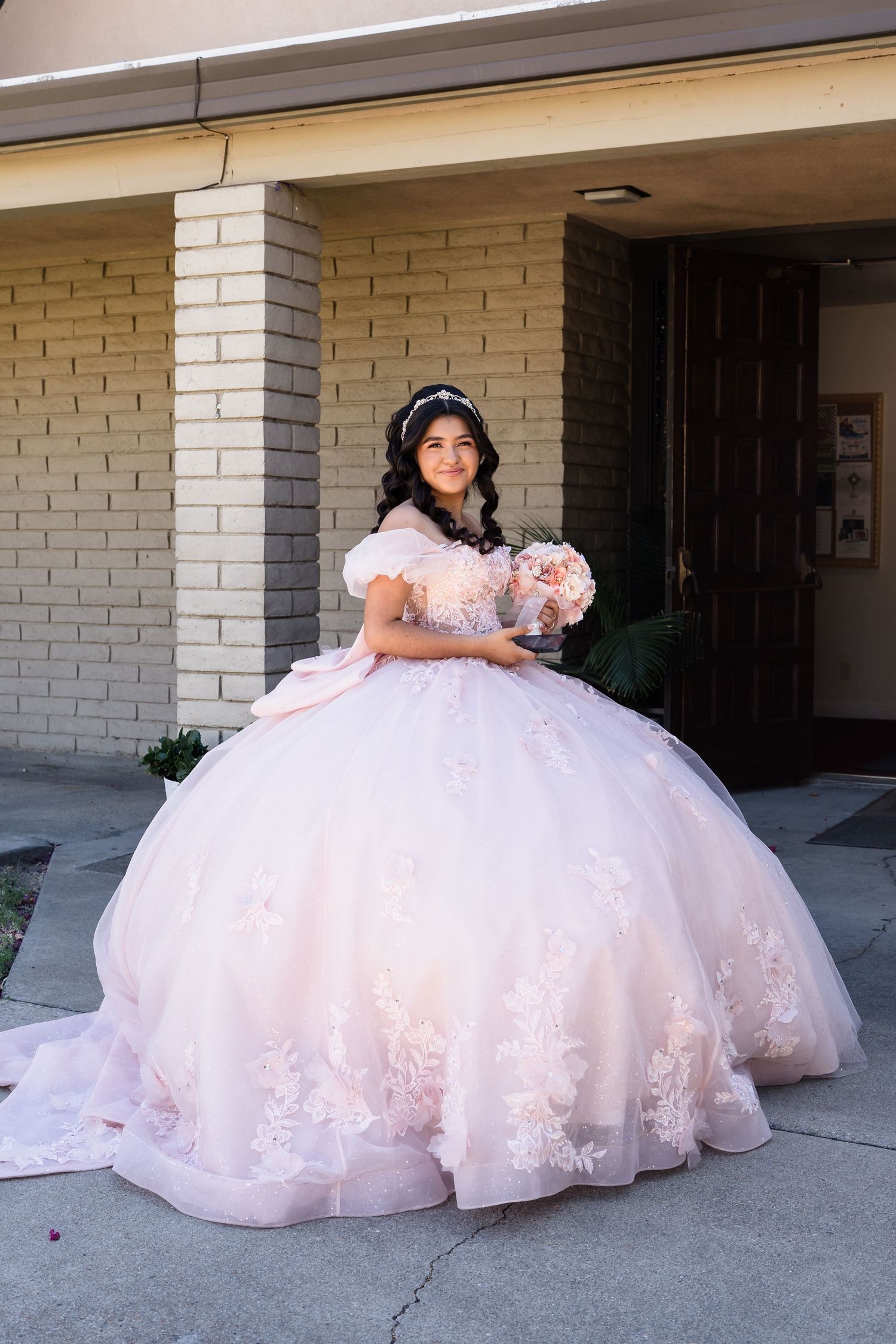 A young Quinceañera poses in a voluminous pink ball gown with lace, holding a bouquet outside.