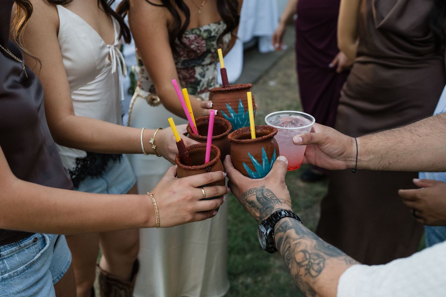 Group of friends toasting with colorful drinks in clay and plastic cups at an outdoor celebration.