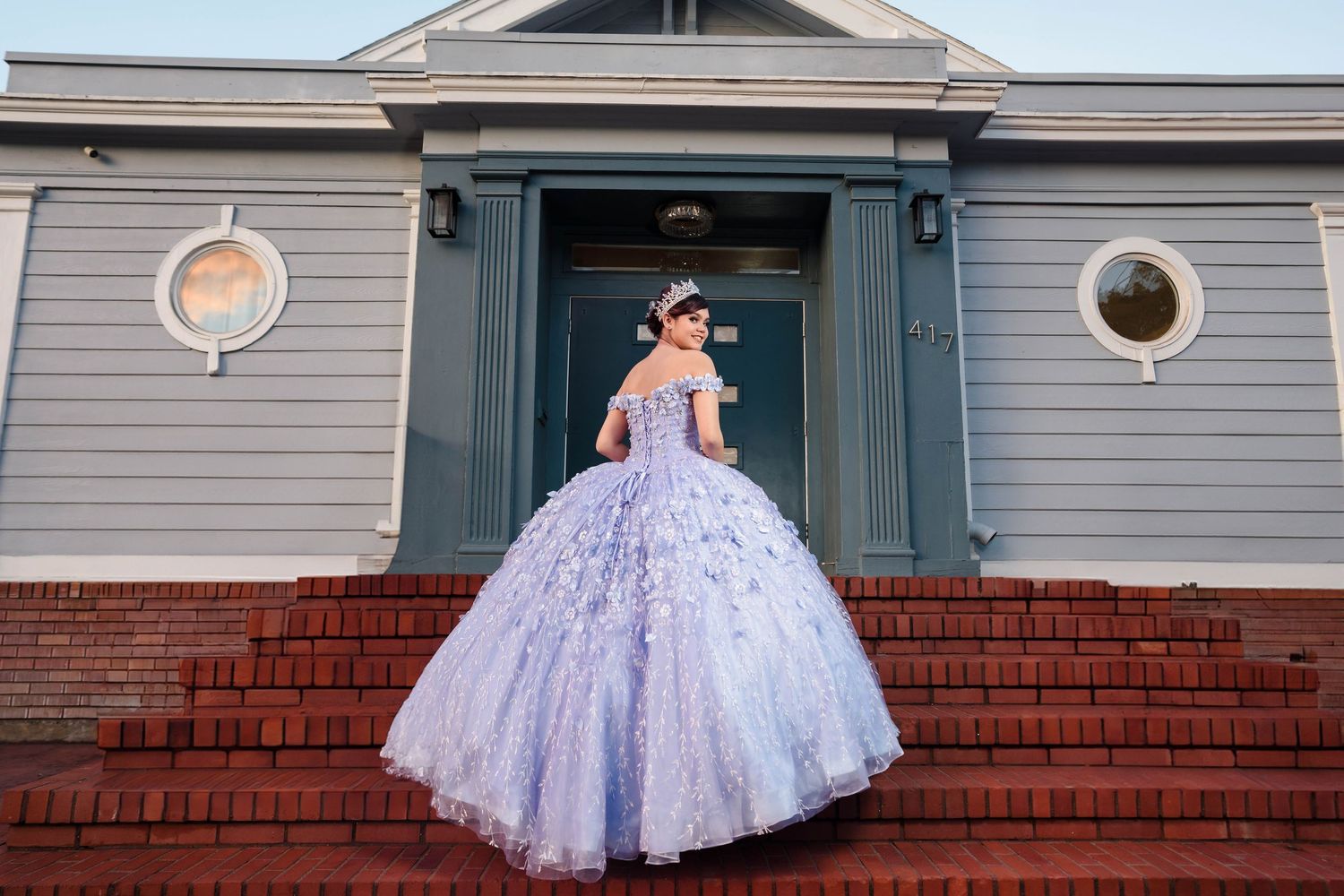 Young woman in lavender ball gown with floral details poses on brick steps outside a teal building.