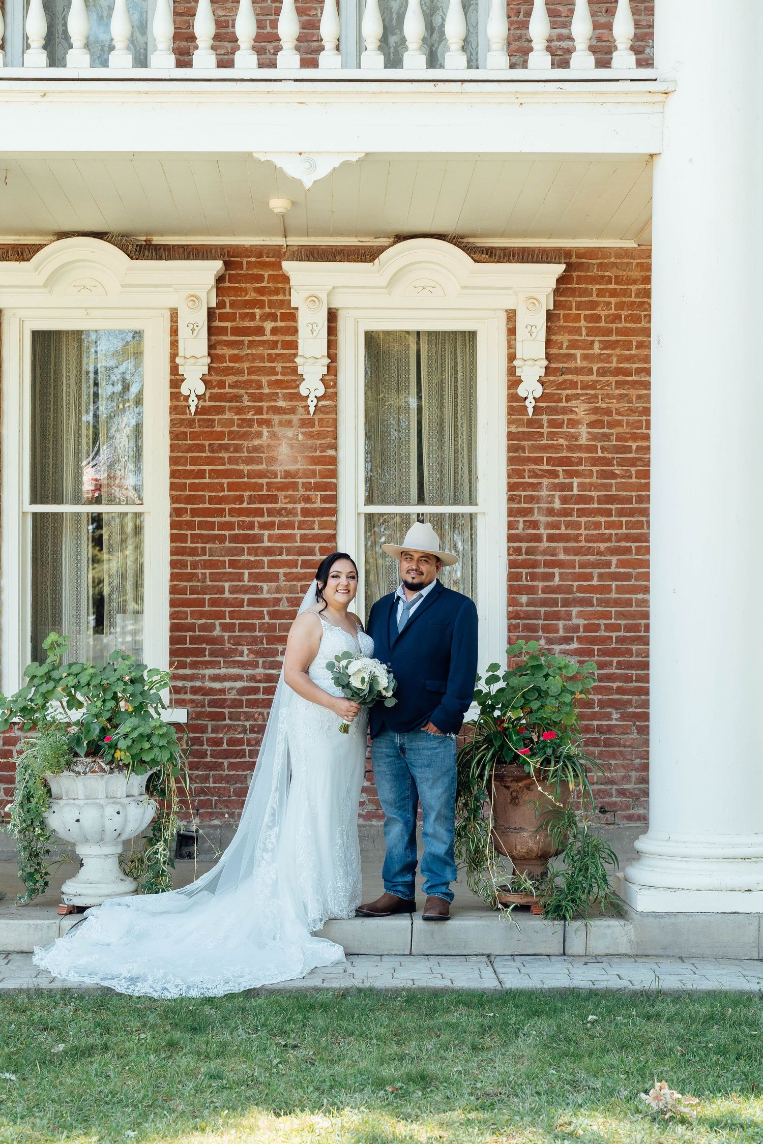 Smiling bride and groom pose on steps of elegant brick venue with white trim and lush planters.