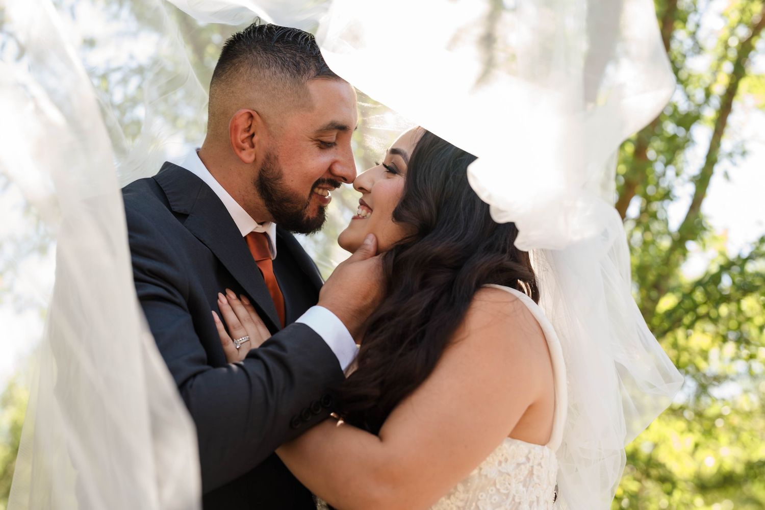 A bride and groom share a romantic moment under a flowing white veil outdoors on their wedding day.