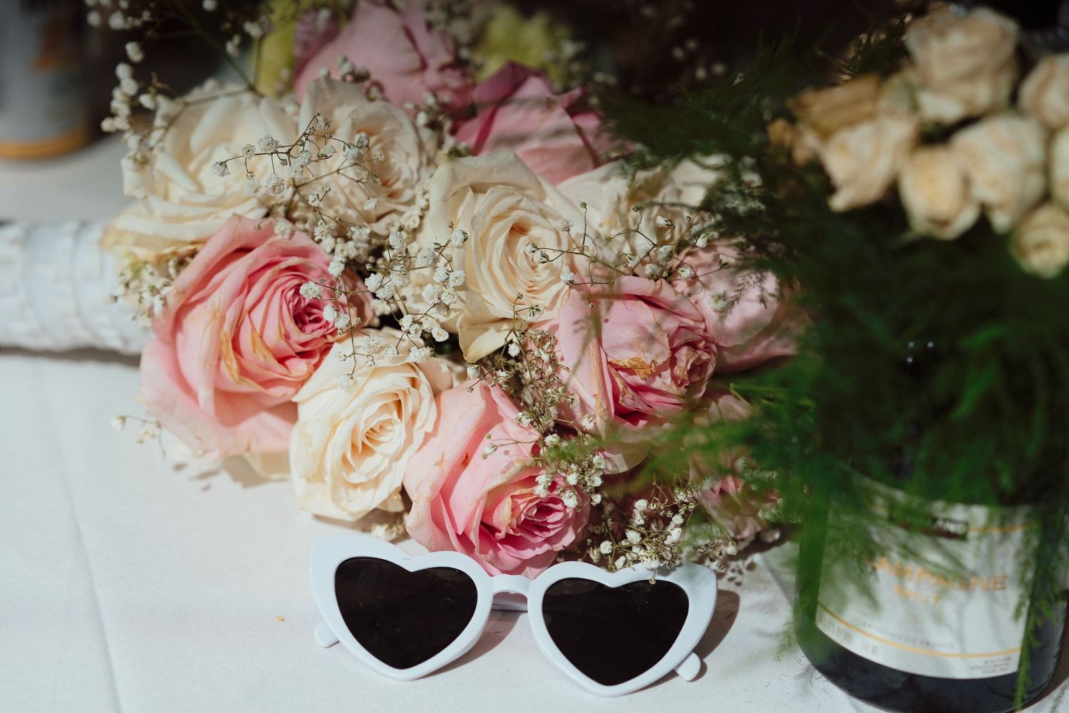 Pink and cream roses bouquet with baby's breath next to white heart-shaped sunglasses on a white table.