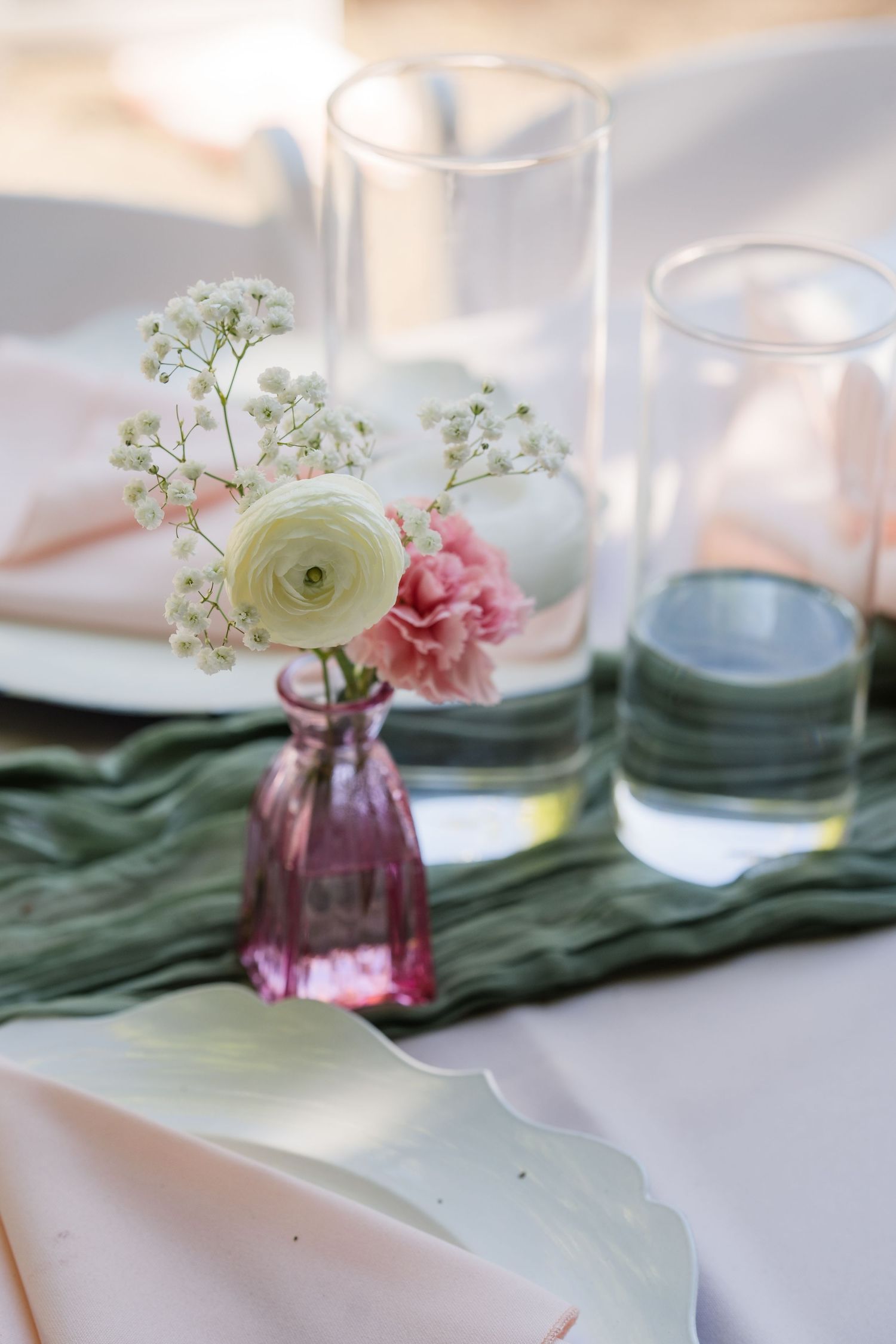 Elegant wedding table with pink ranunculus and baby's breath in glass vase on green runner.