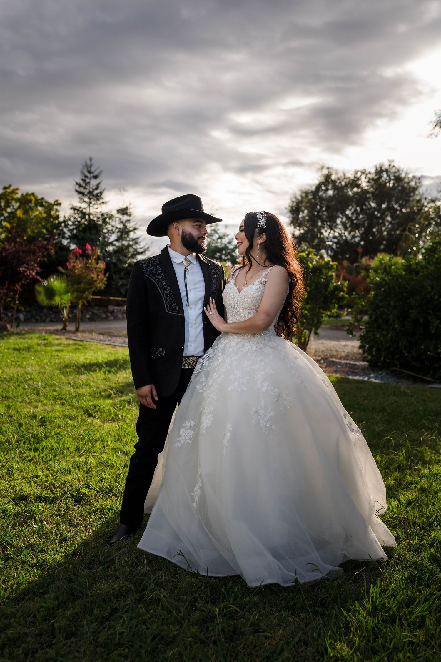 Bride in white ball gown and groom in black charro suit with cowboy hat share a romantic moment in a lush green garden.