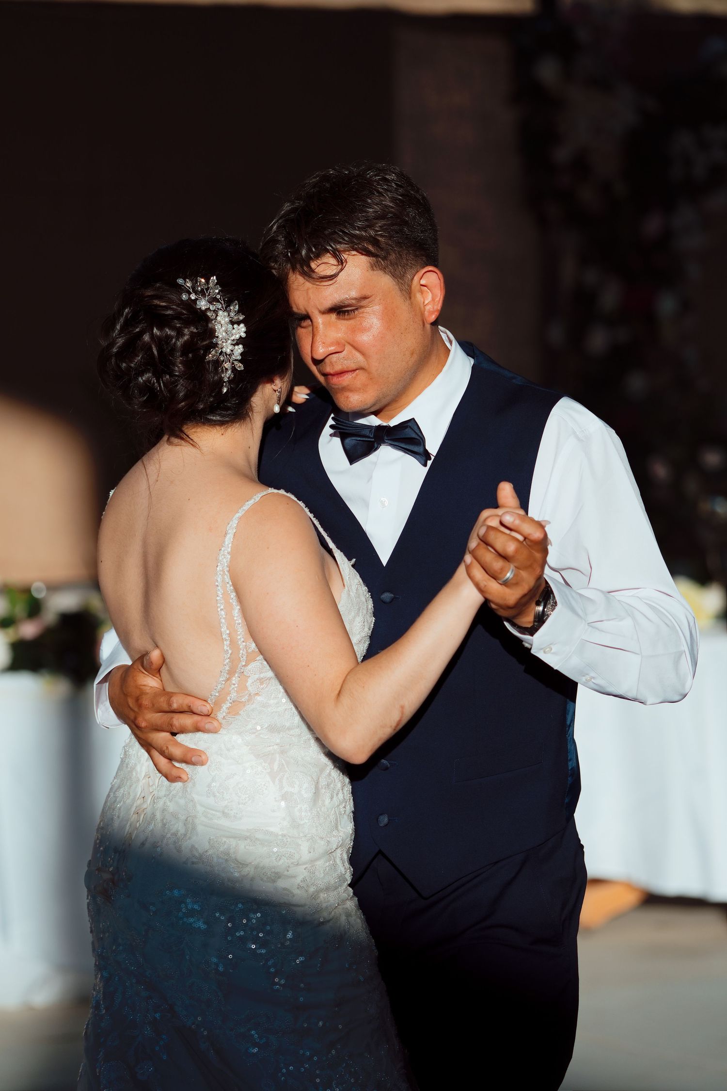 Bride and groom share emotional first dance, groom in navy vest and bow tie holds bride in lace gown.