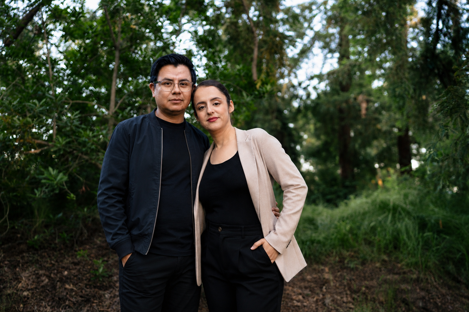 Couple stands together in a lush green forest, man in black jacket and woman in beige blazer.