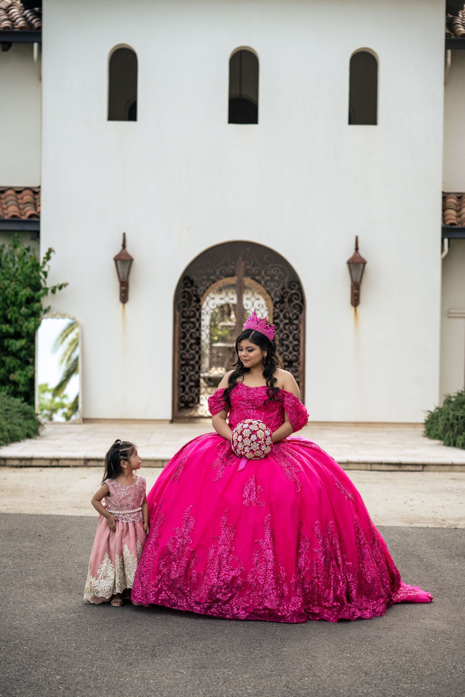 Quinceañera in fuchsia gown holds bouquet while young girl in pink dress stands beside her.