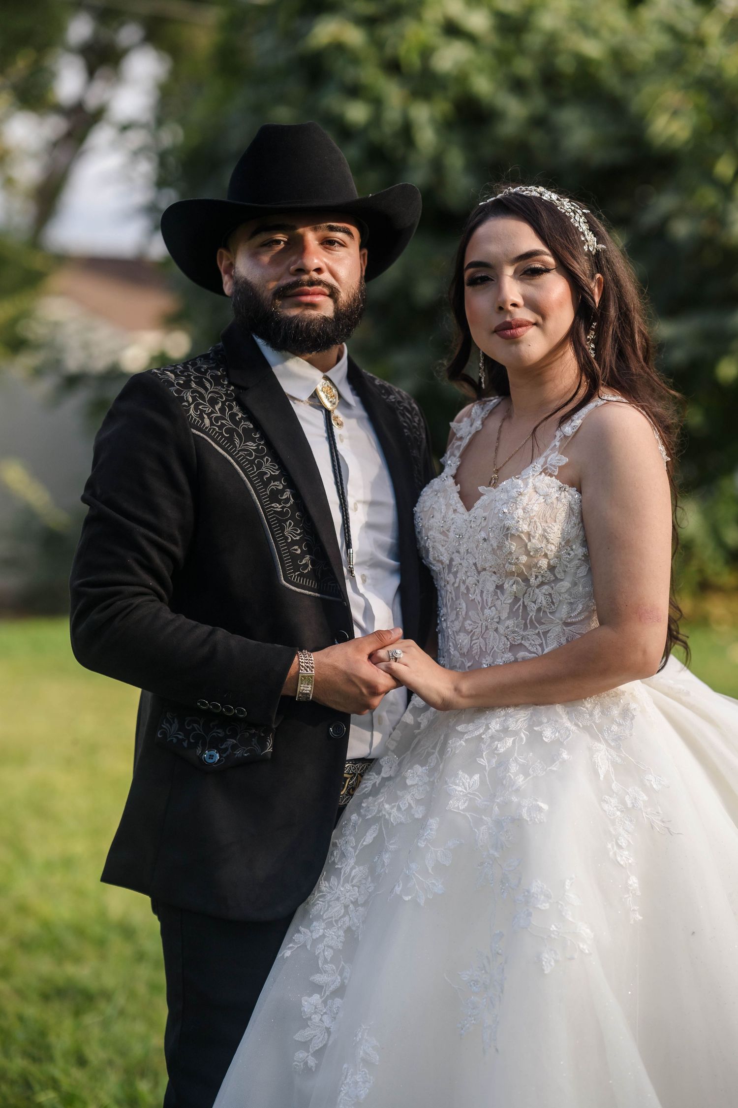Elegant couple poses outdoors, groom in black embroidered suit and cowboy hat, bride in lace gown.