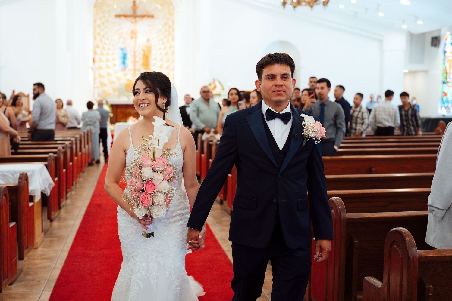 Bride and groom walk down church aisle after wedding ceremony, bride holding pink bouquet, groom in navy suit.