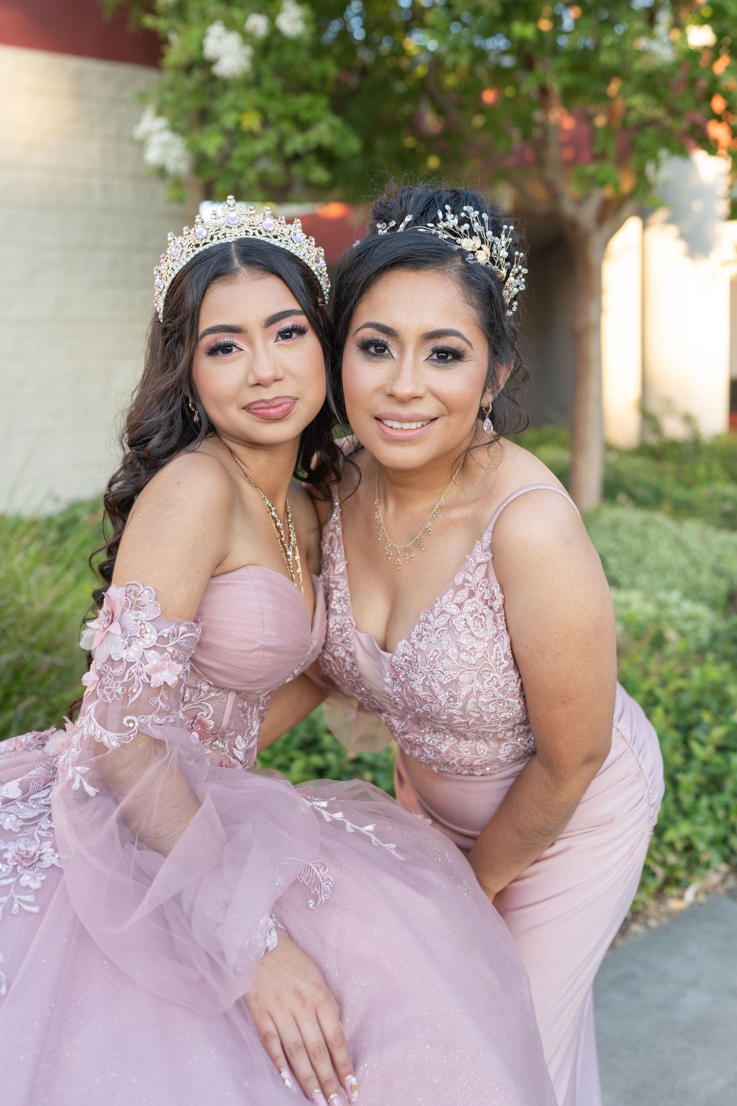 Quinceañera girl wearing crown poses with woman in matching pink dress outdoors.