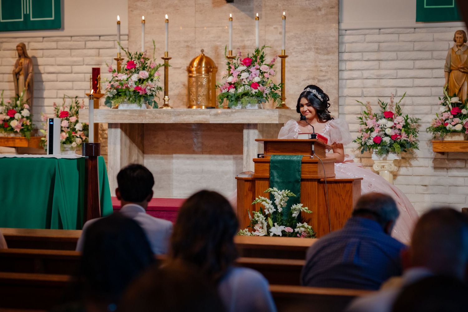 Woman in elegant attire speaks at a church altar adorned with floral arrangements during a religious ceremony.