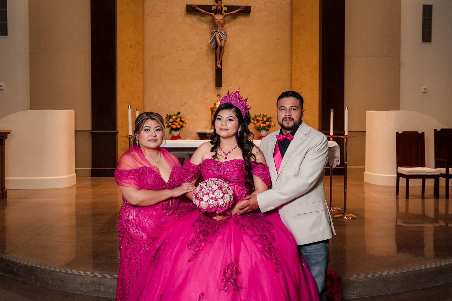 Quinceañera girl in pink gown holding bouquet posing with parents at church altar.