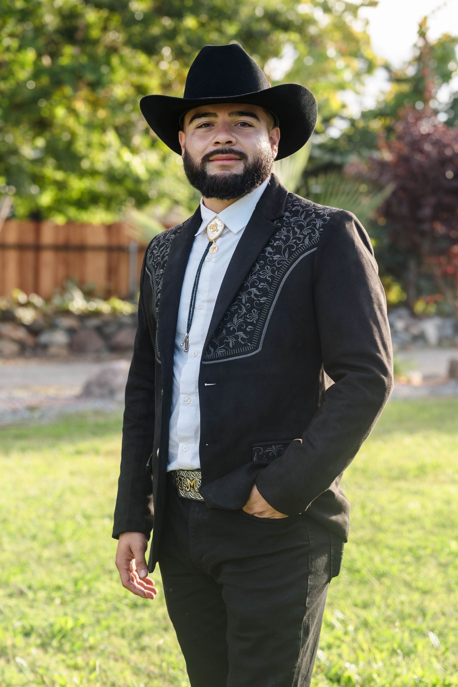 Stylish groom in black embroidered charro suit and black cowboy hat posing outdoors in garden.