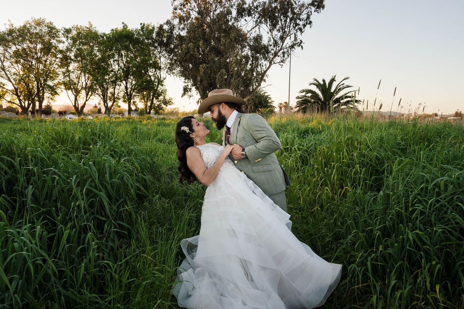 Bride and groom kissing in a lush green field at sunset, bride in white ball gown, groom in suit and cowboy hat.