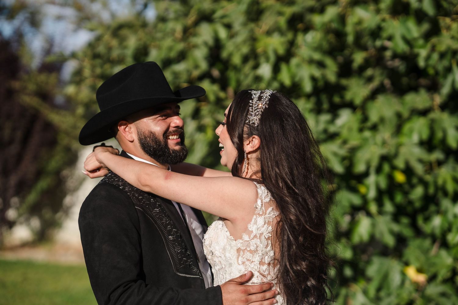 A smiling bride and groom in wedding attire embrace outdoors, groom wearing a black cowboy hat, surrounded by lush greenery.