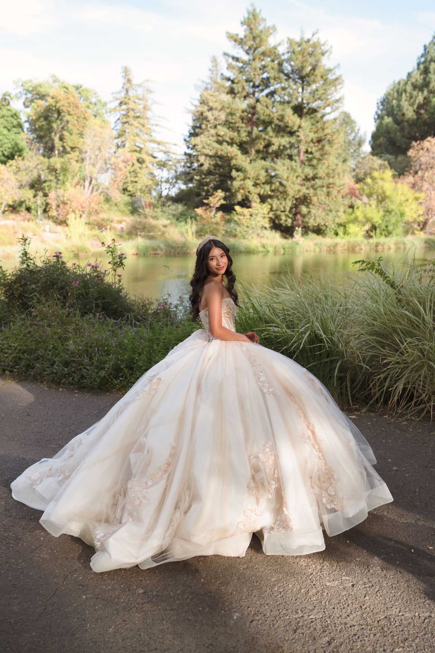 Young woman in a flowing champagne ball gown with floral lace details poses gracefully by a serene park pond.