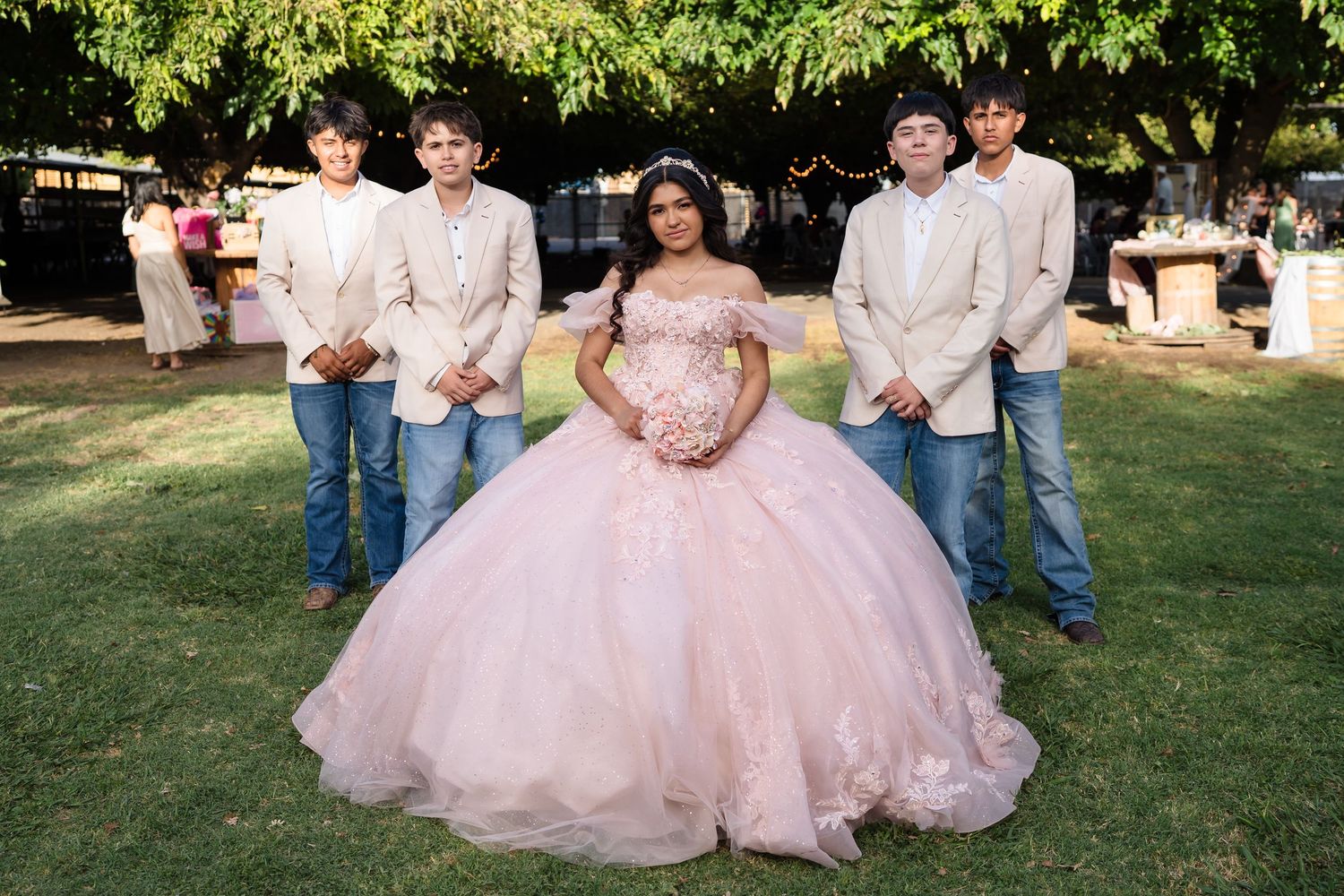 Young woman in pink quinceañera ball gown poses with four young men in beige suits and jeans outdoors.
