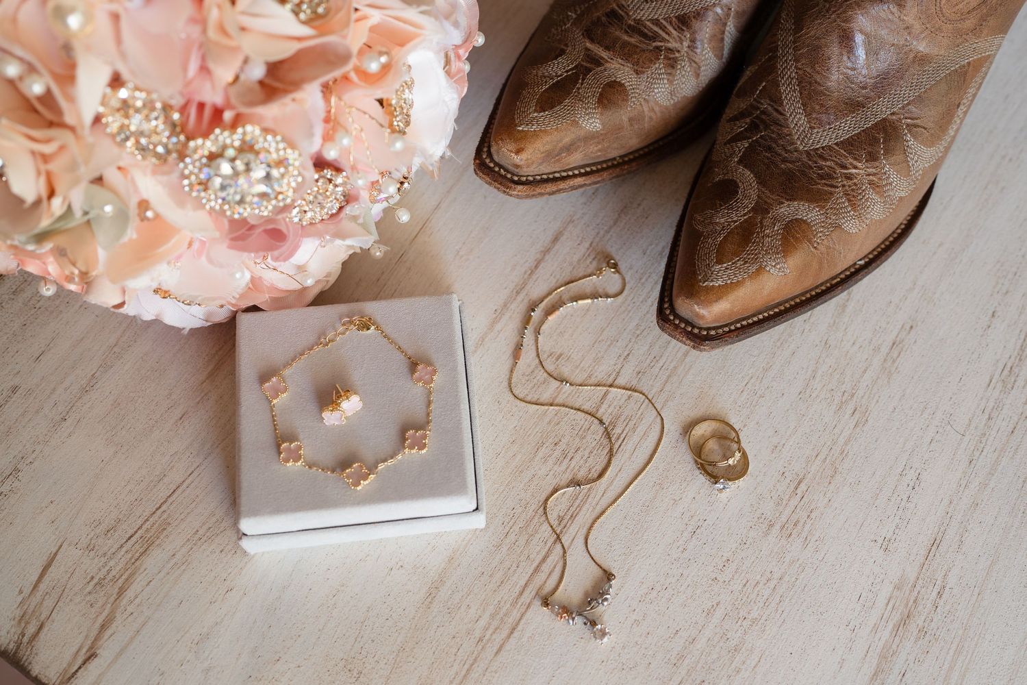 Rustic wedding flat lay with pink floral bouquet, gold jewelry set, delicate necklace, ring, and brown cowboy boots on wood.