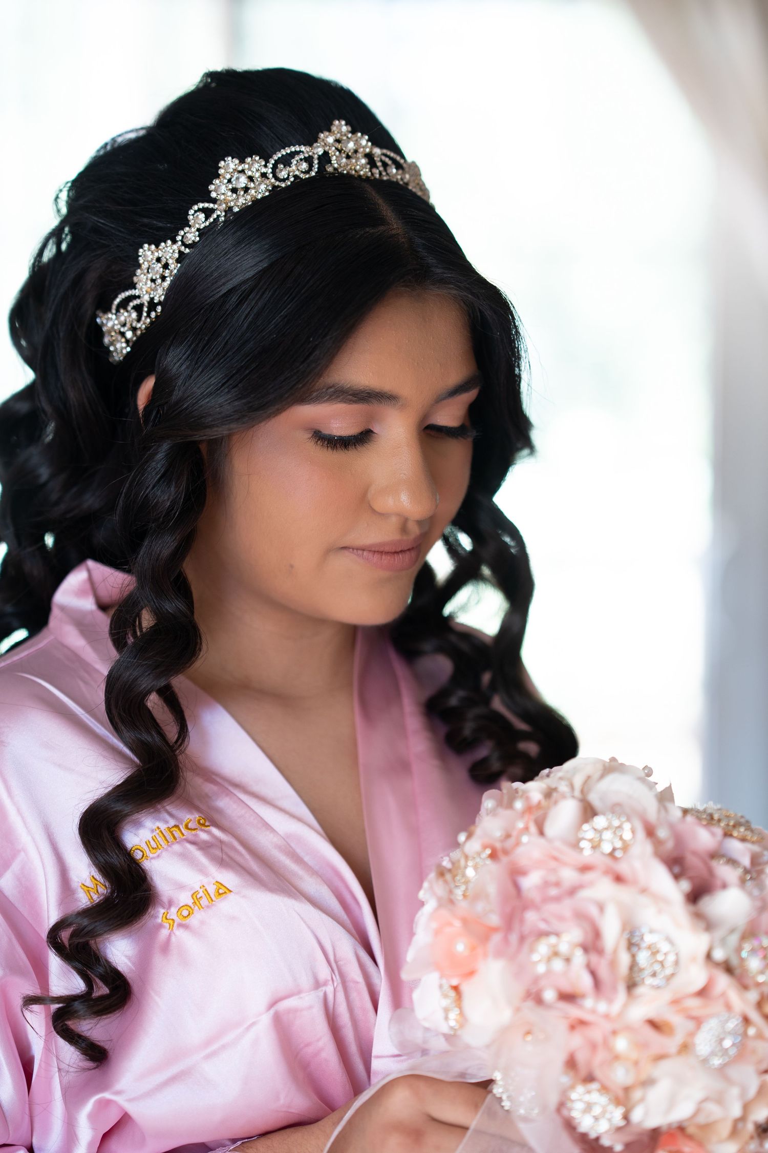 Young Latina quinceañera in pink satin robe wearing a crystal tiara holds a blush pink floral bouquet.